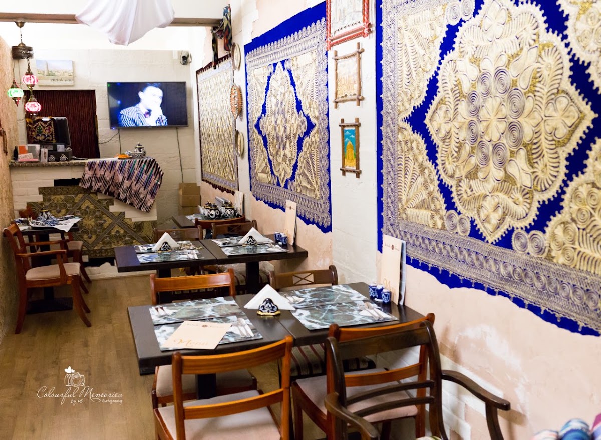 Inside Uzbek Corner: dark wooden tables laid with cutlery, napkins and small blue-and-white teapots, beneath large gold-embroidered suzani hangings on a royal blue ground. Coloured glass lamps hang by the wall and the floor is pale oak.