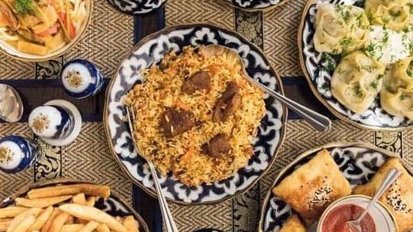 A table seen from above: a platter of rice and lamb in the centre, dumplings in a creamy sauce, pastries with red sauce, salad, chips and small blue-and-white lidded pots.